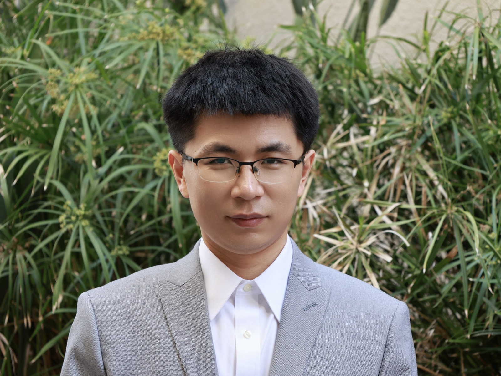 Portrait of Jinyu Gao, wearing glasses and a light gray jacket, standing in front of green plants.